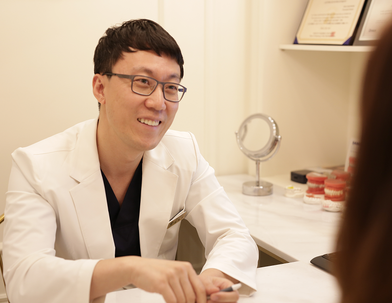 흰 가운을 입은 원장이 상담 데스크에 앉아 마주 앉은 환자를 향해 이야기하는 모습, 옆에는 거울과 치아 모형이 놓여 있음