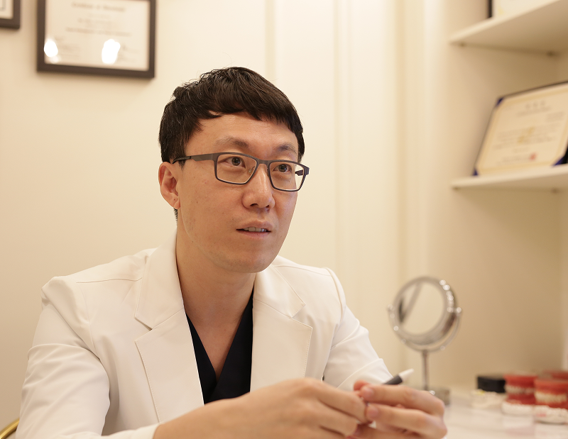 흰 가운을 입은 원장이 손거울과 치아 모형이 놓인 상담 데스크에서 펜을 들고 설명하는 모습